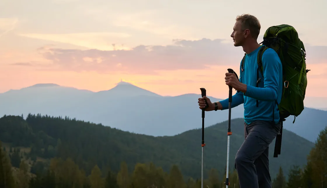 Wanderer mit Ausrüstung auf einer Kuppe, vor sich blau schimmernde Bergkämme im Abendlicht